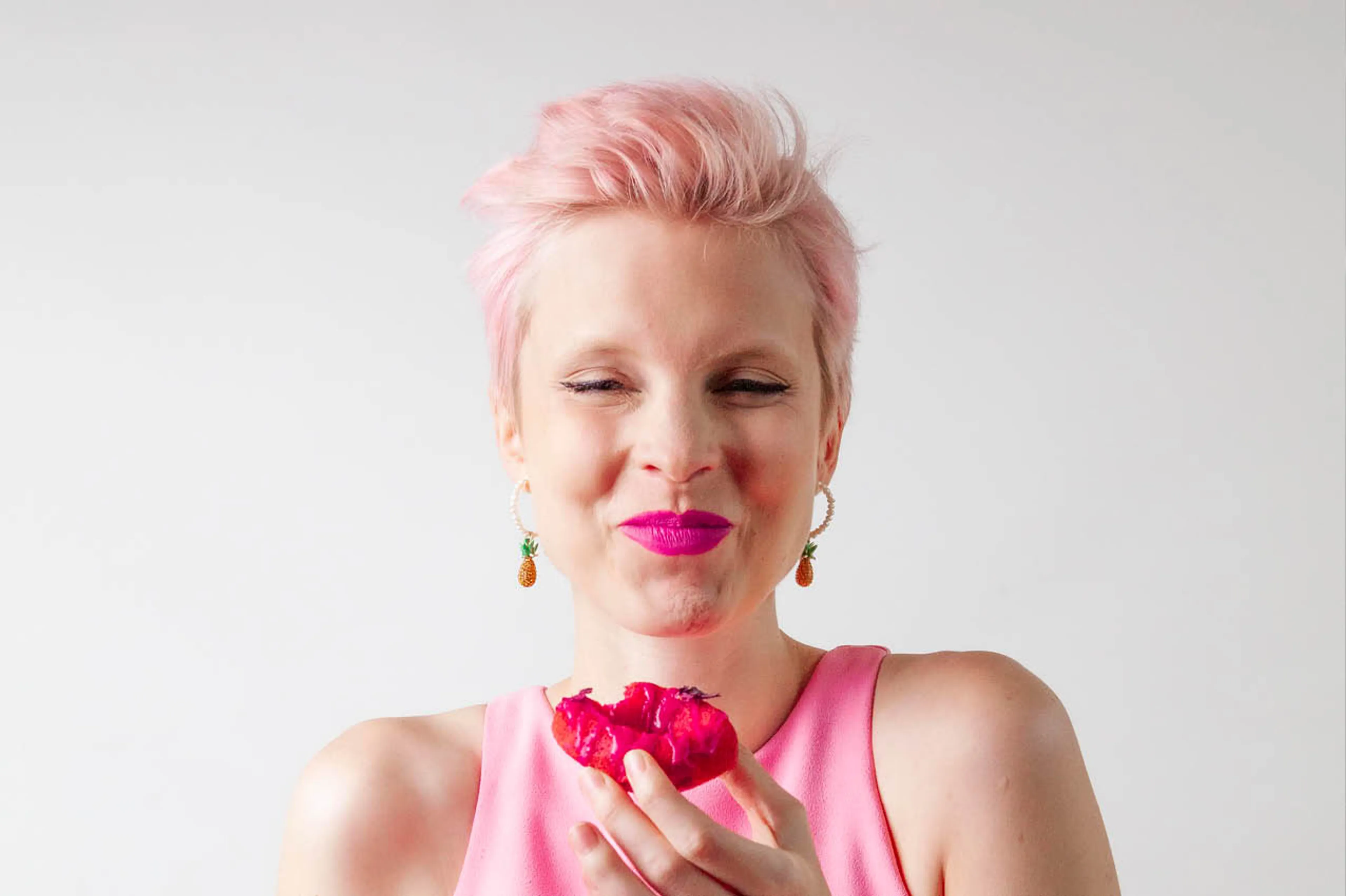 Eveline van Vegamuze met kort roze stekeltjeshaar, felroze lippen en in haar hand een donut van rode biet. Ze draagt een lichtroze tanktop en lacht met geknepen ogen naar de camera